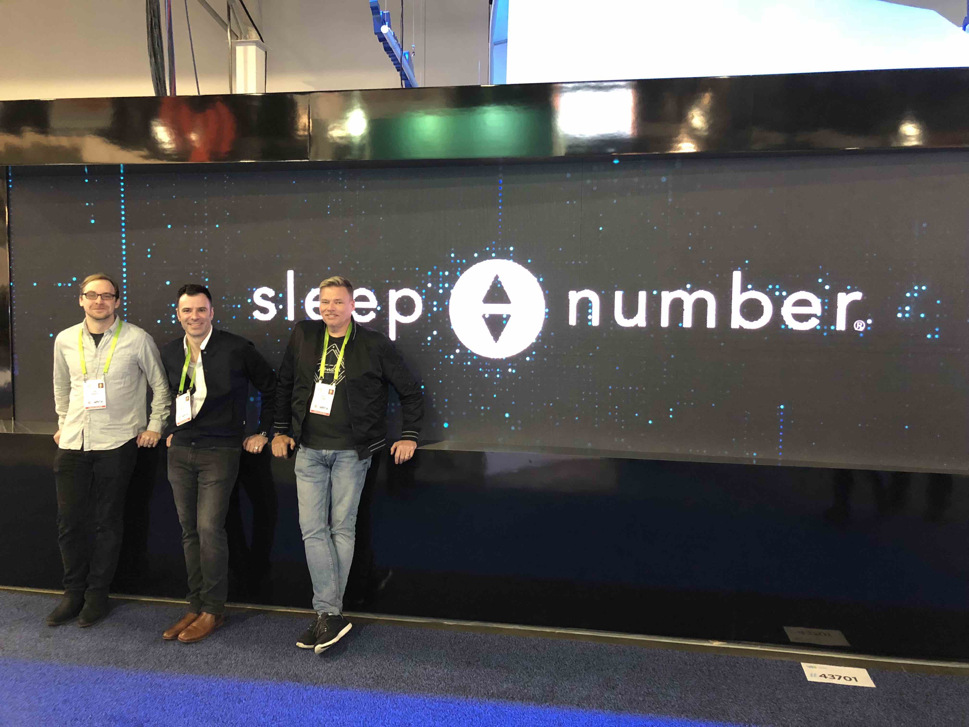 Three men standing in front of a digital screen reading "Sleep Number" at CES trade show