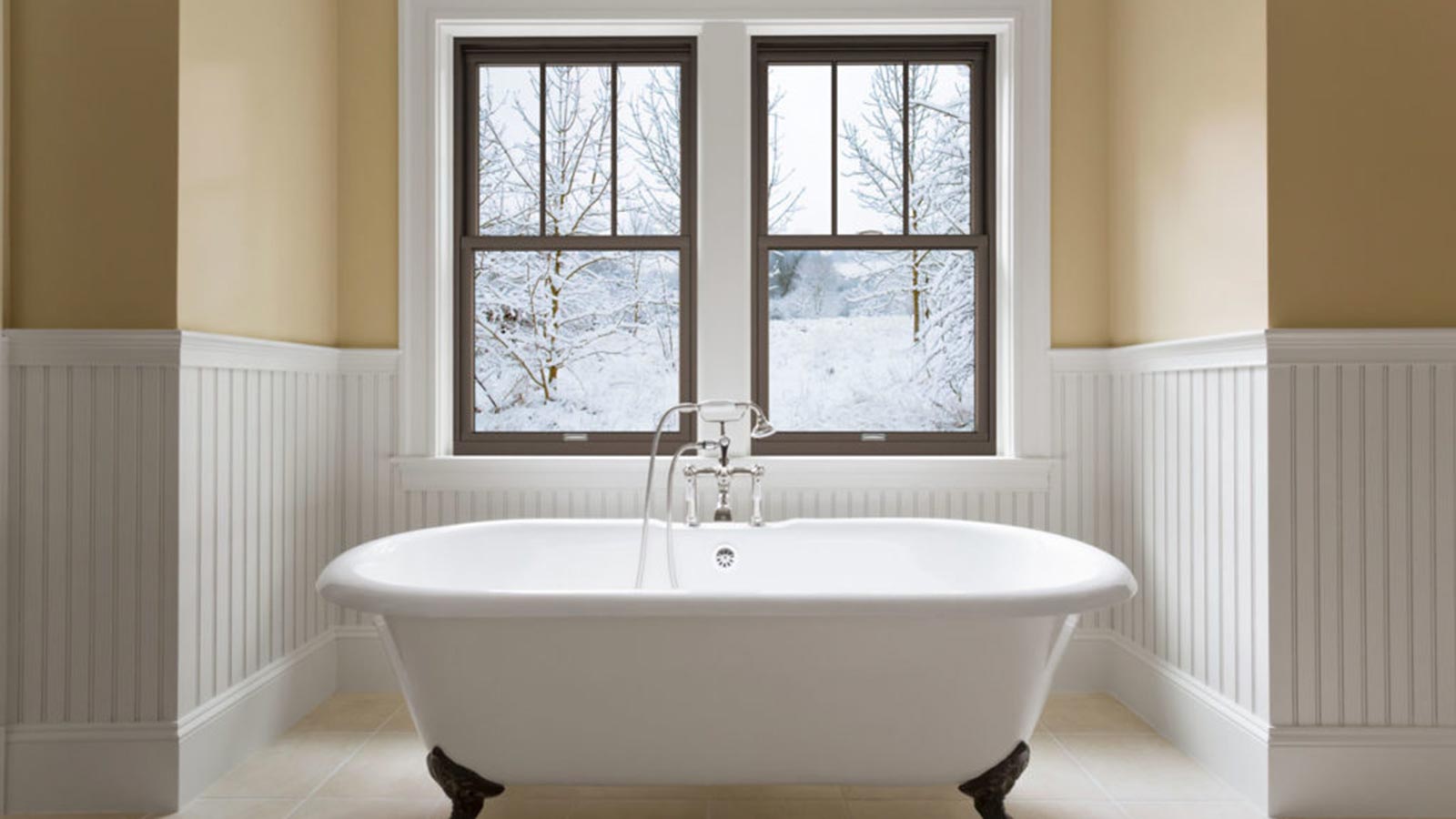 Claw-foot bathtub in front of large glass windows overlooking a snowy backyard